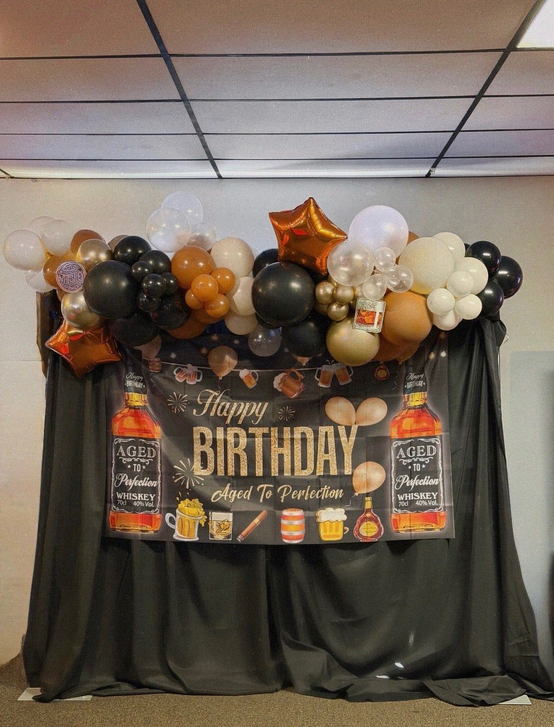 A black, gold, orange and clear balloon garland across the top of an aged-to-perfection birthday backdrop