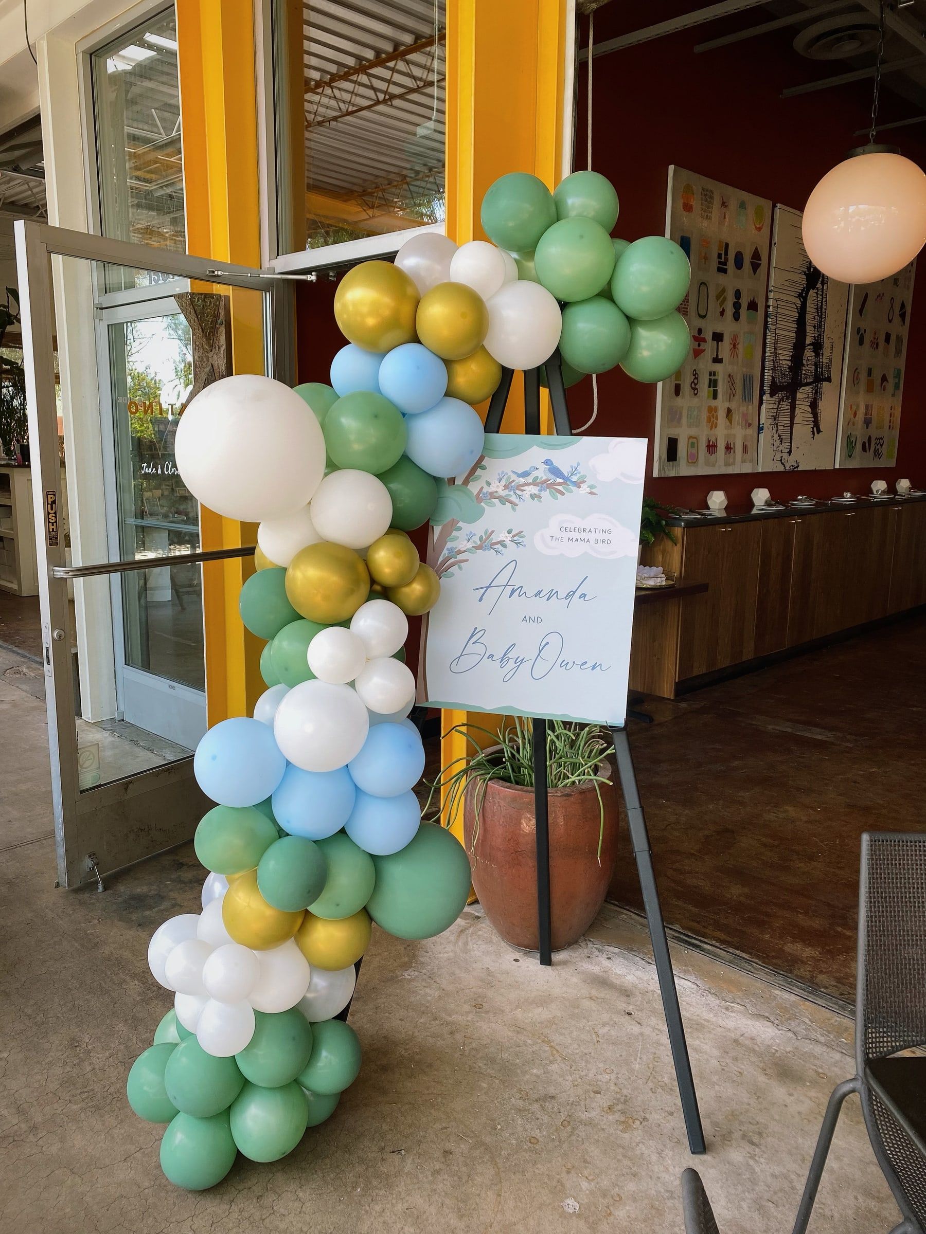 An organic balloon column in sage, sky blue, white and gold rising beside the open door of a venue, next to a baby shower welcome sign on an easel