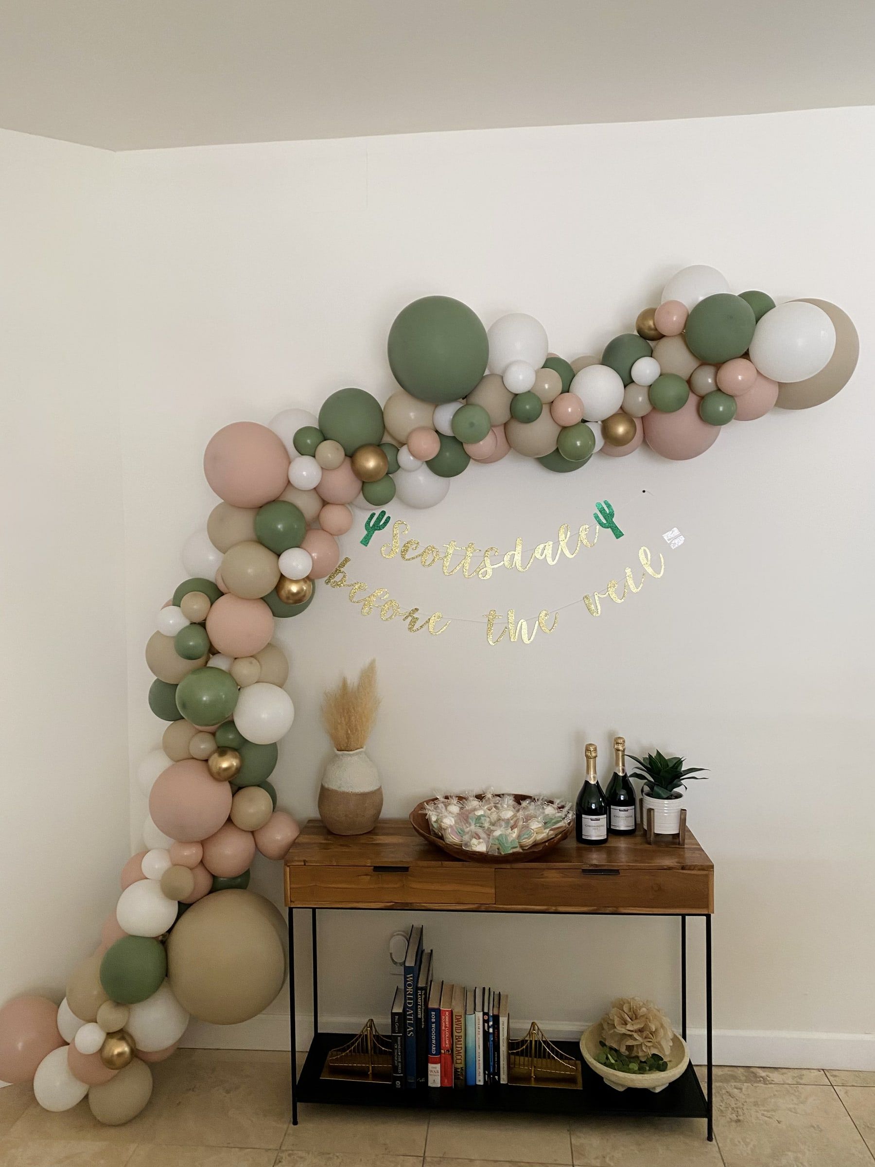 An organic balloon garland in blush, sage, cream and gold sweeping from the floor up and across a wall above a console table