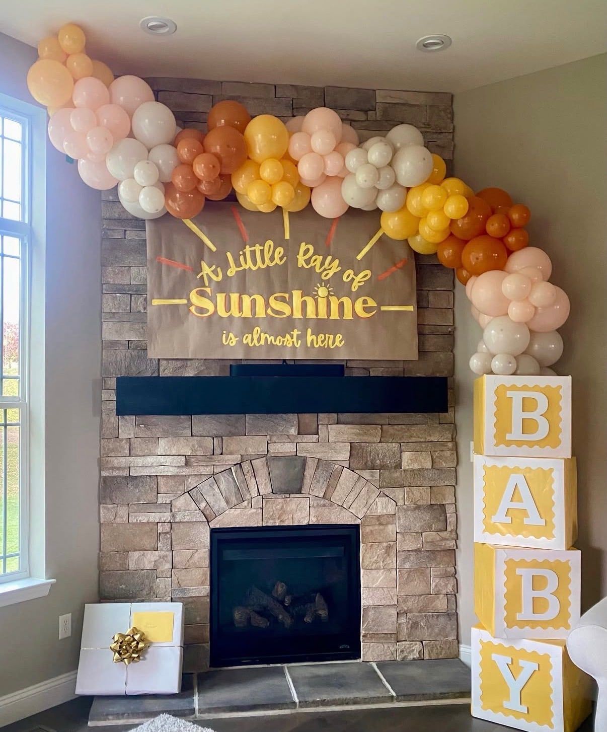 A cream, peach and orange balloon arch over a stone fireplace with a sunshine sign and BABY blocks