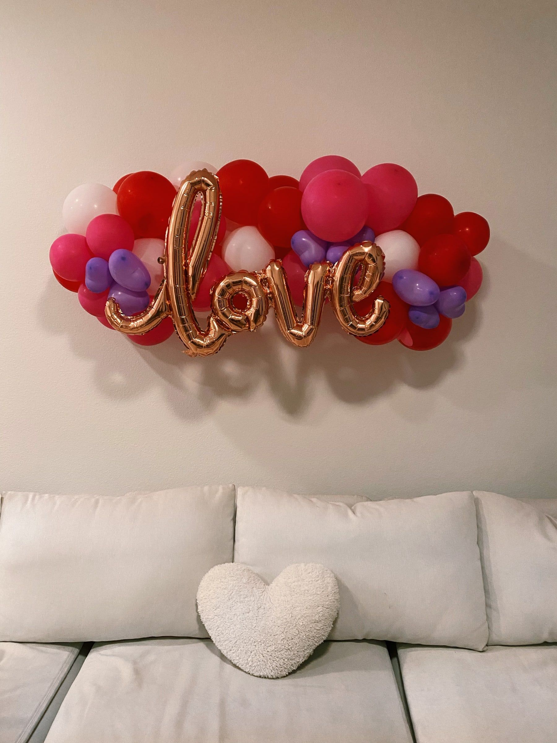 A red, pink, white and lilac balloon garland on a living room wall with rose gold script "love" lettering, above a white sofa