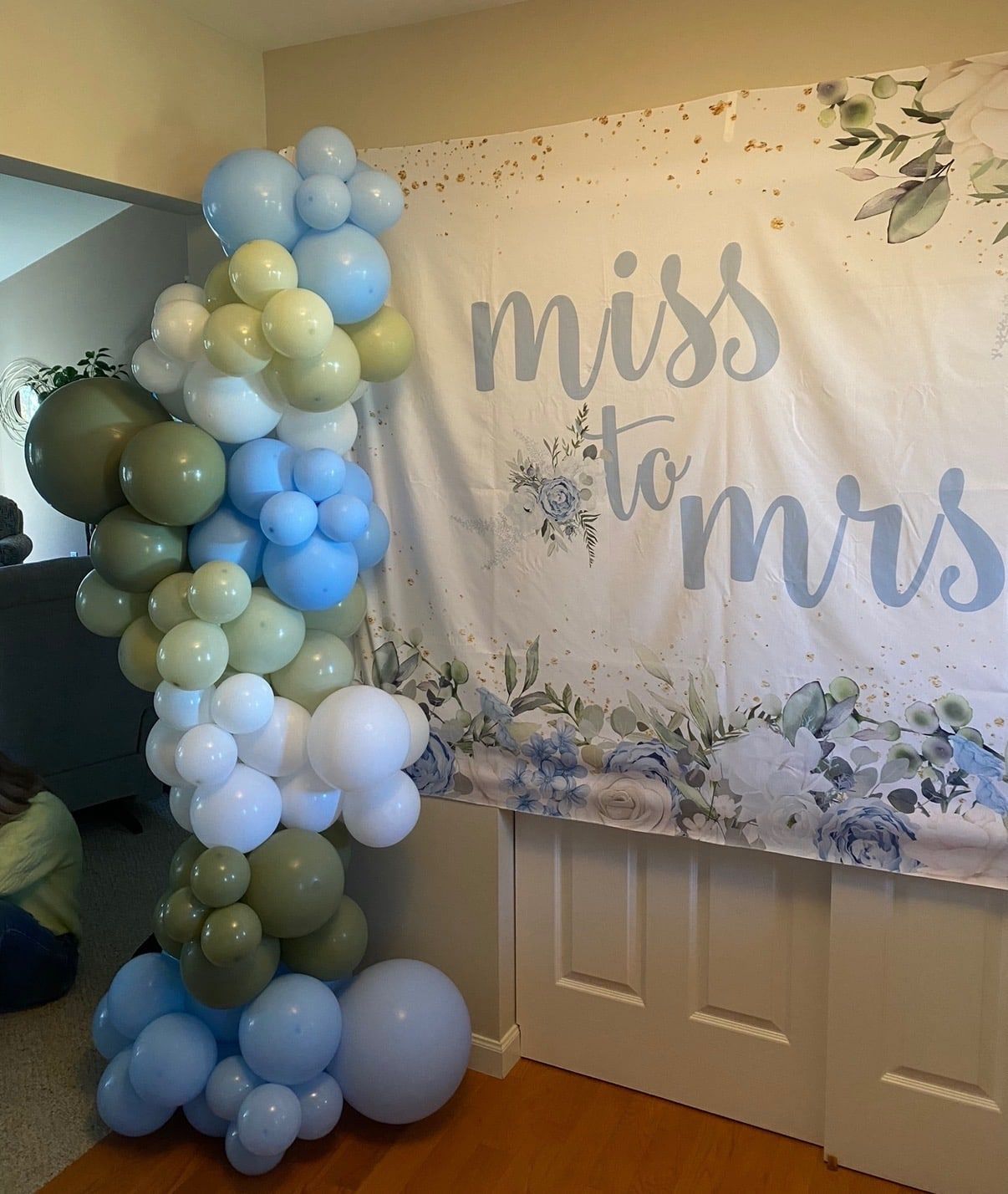 An organic column of blue, sage and white balloons beside a Miss to Mrs backdrop
