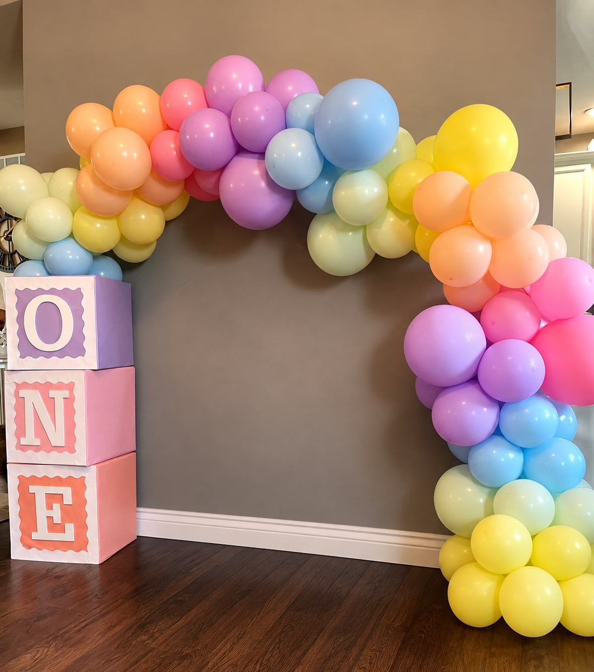 A large organic arch of pastel rainbow balloons beside stacked O, N and E letter blocks