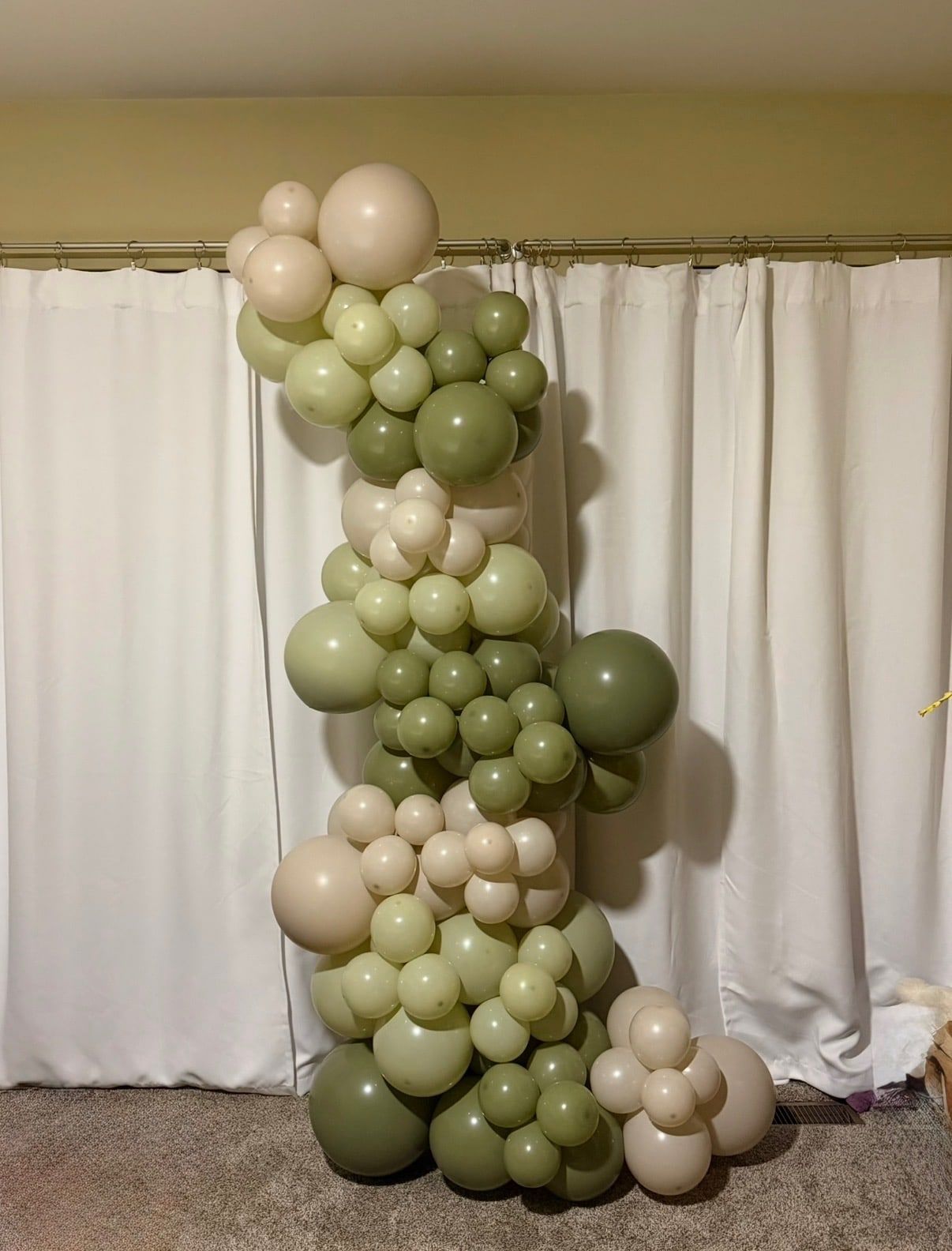 A tall organic column of sage green and cream balloons against white curtains