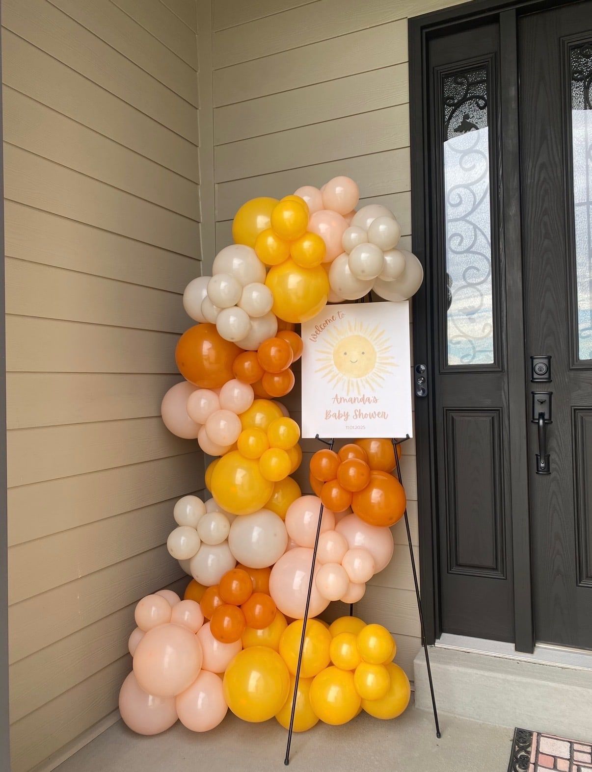An organic balloon column in yellow, orange, peach and cream rising beside a front door, with a sunshine welcome sign on an easel